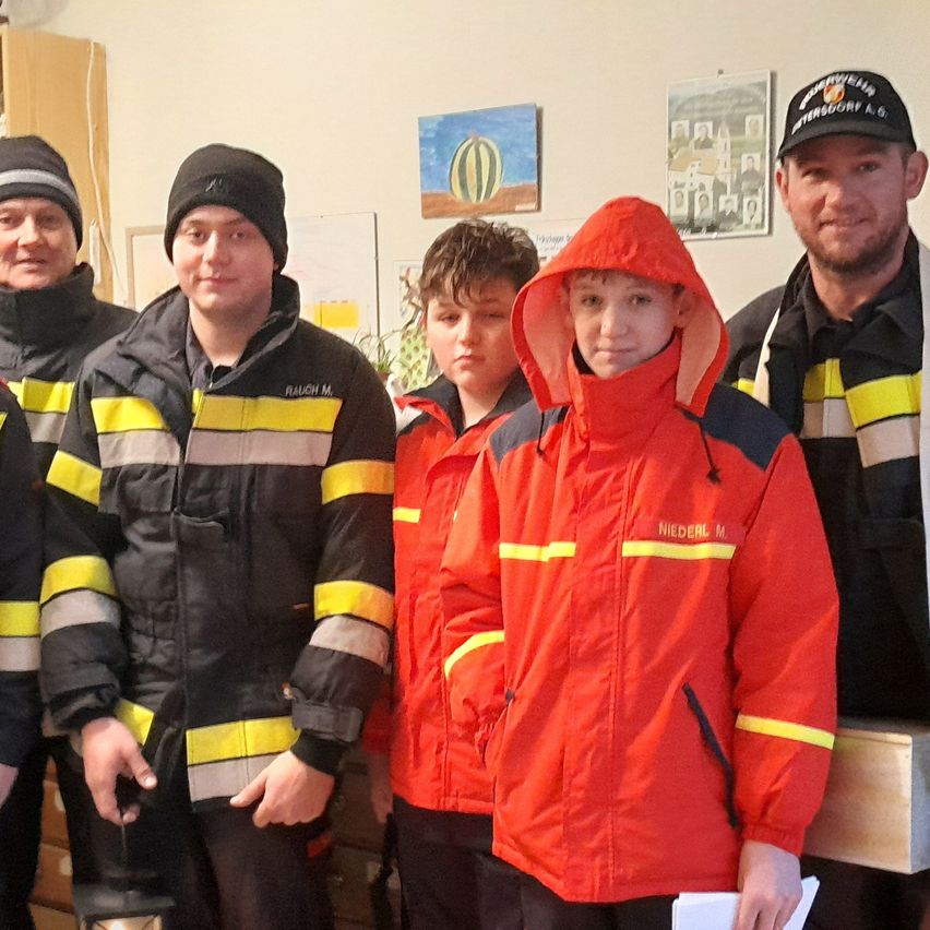 Eine Gruppe Feuerwehrleute in roten Jacken mit gelben Streifen und schwarzen Mützen posiert für ein Foto in einem Raum mit Gemälden an der Wand.