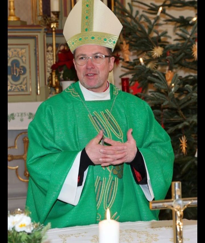 Ein Mann in grüner und weißer Priesterrobe steht vor einem Weihnachtsbaum und spricht. Er trägt eine Brille und einen Ring. Hinter ihm befindet sich eine Wand mit einem Emblem und einer Kerze.