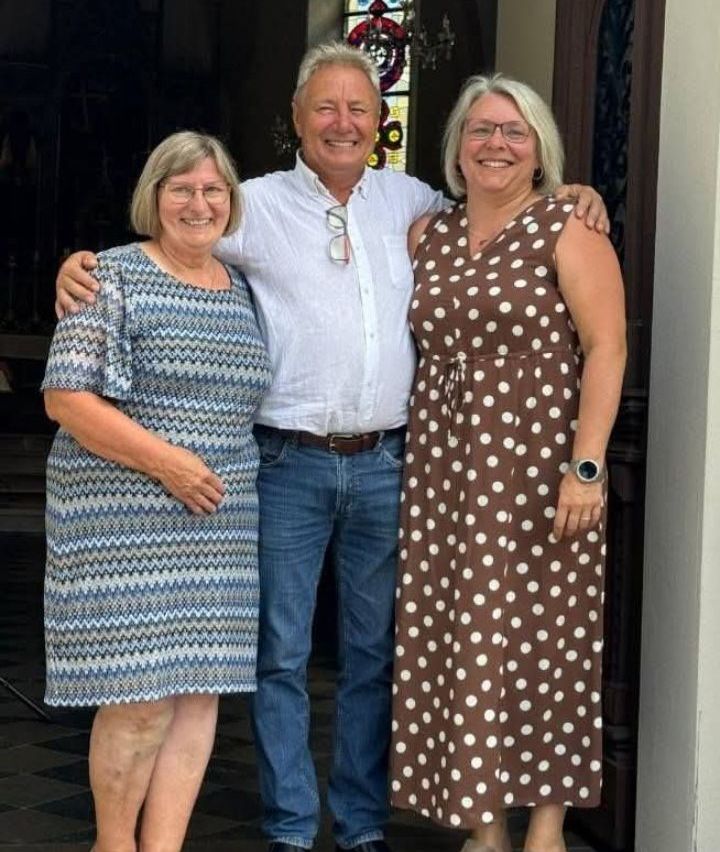 Drei Menschen, zwei Frauen und ein Mann, stehen eng beieinander und lächeln für ein Foto vor einem Gebäude.
