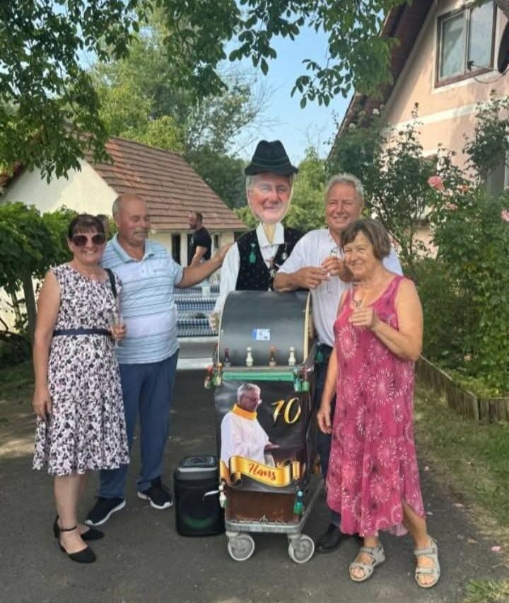 Fünf Personen stehen vor einem Haus, zwei Frauen und drei Männer, einer der Männer ist als Karikatur eines Mannes mit einem Bierfass auf einem Karren verkleidet.