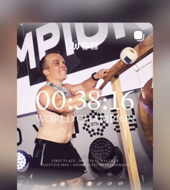 Ein Mann feiert seinen Sieg bei der IMPU1-Meisterschaft. Er hält einen Hammer und lächelt, wobei die Zeit 00:38:16 angezeigt wird. Er trägt eine Medaille und steht vor einem karierten Hintergrund.