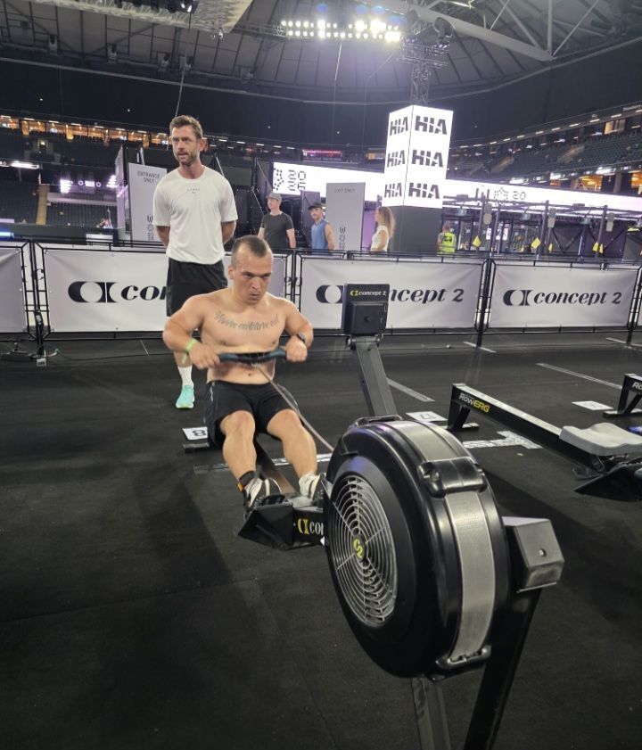 Ein Mann rudert auf einem Rudergerät bei einer Indoor-Veranstaltung. Ein Mann in einem weißen T-Shirt steht hinter ihm. Der Bereich hat Banner mit dem Text 'Concept 2'.