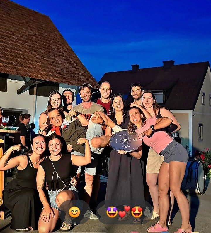 Eine Gruppe von Menschen, einige halten eine Plakette, stehen vor einem Haus unter einem blauen Himmel und lächeln für ein Foto.