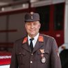 Ein Mann in Uniform steht vor einem Feuerwehrauto. Er trägt eine Mütze, eine Medaille und eine Krawatte. Er lächelt und posiert wahrscheinlich für ein Foto.