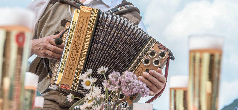 Ein Mann in traditioneller Kleidung spielt Akkordeon mit Blumen und Biergläsern im Hintergrund.