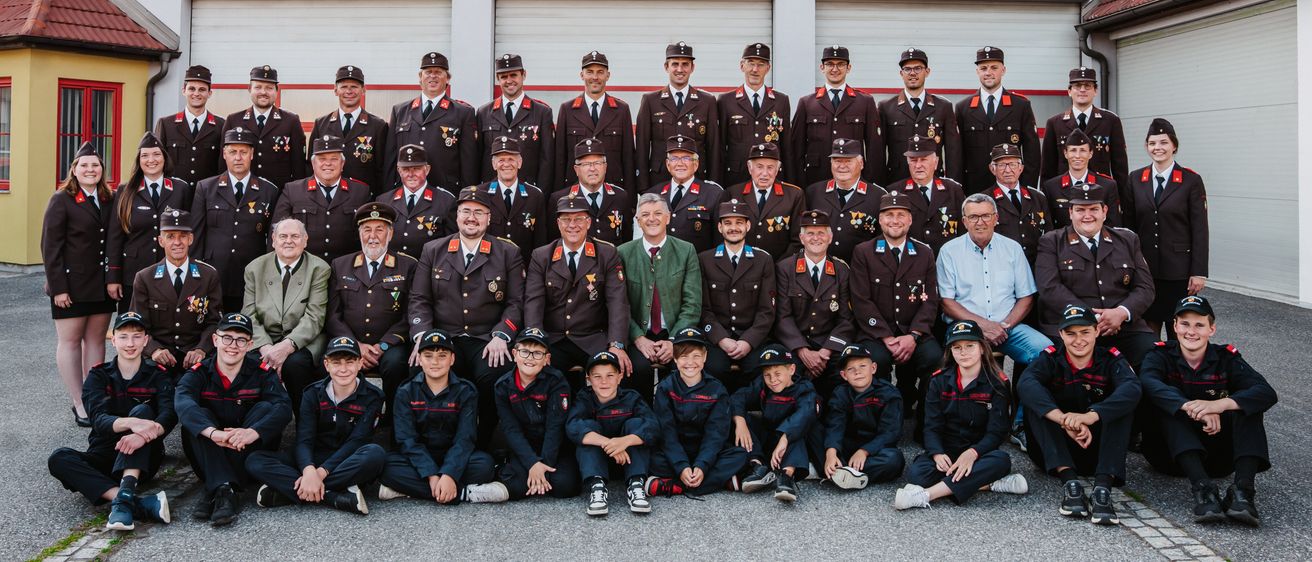Ein Gruppenfoto von Feuerwehrleuten in Uniform, einschließlich Männer und Kinder, steht vor einem Feuerwehrhaus namens 'Feuerwehr Wagendorf.' Sie tragen Mützen und Uniformen mit verschiedenen Abzeichen und Medaillen.