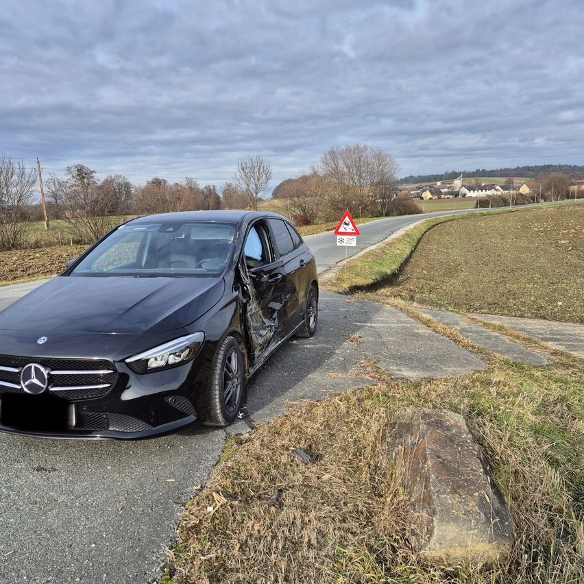 Ein schwarzer Mercedes-Kombi ist am Straßenrand geparkt und vorne beschädigt. Der Himmel ist bewölkt. Ein dreieckiges Warnschild befindet sich in der Nähe.