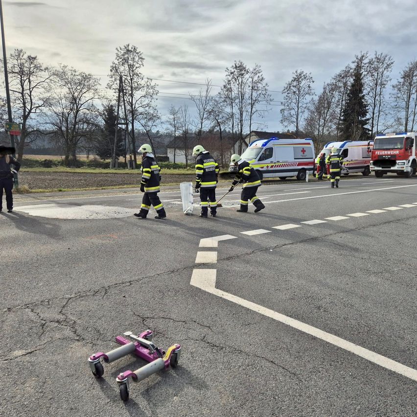 Eine Gruppe von Feuerwehrleuten arbeitet auf der Straße mit einem Wagenheber auf dem Boden. In der Nähe sind auch Krankenwagen geparkt.