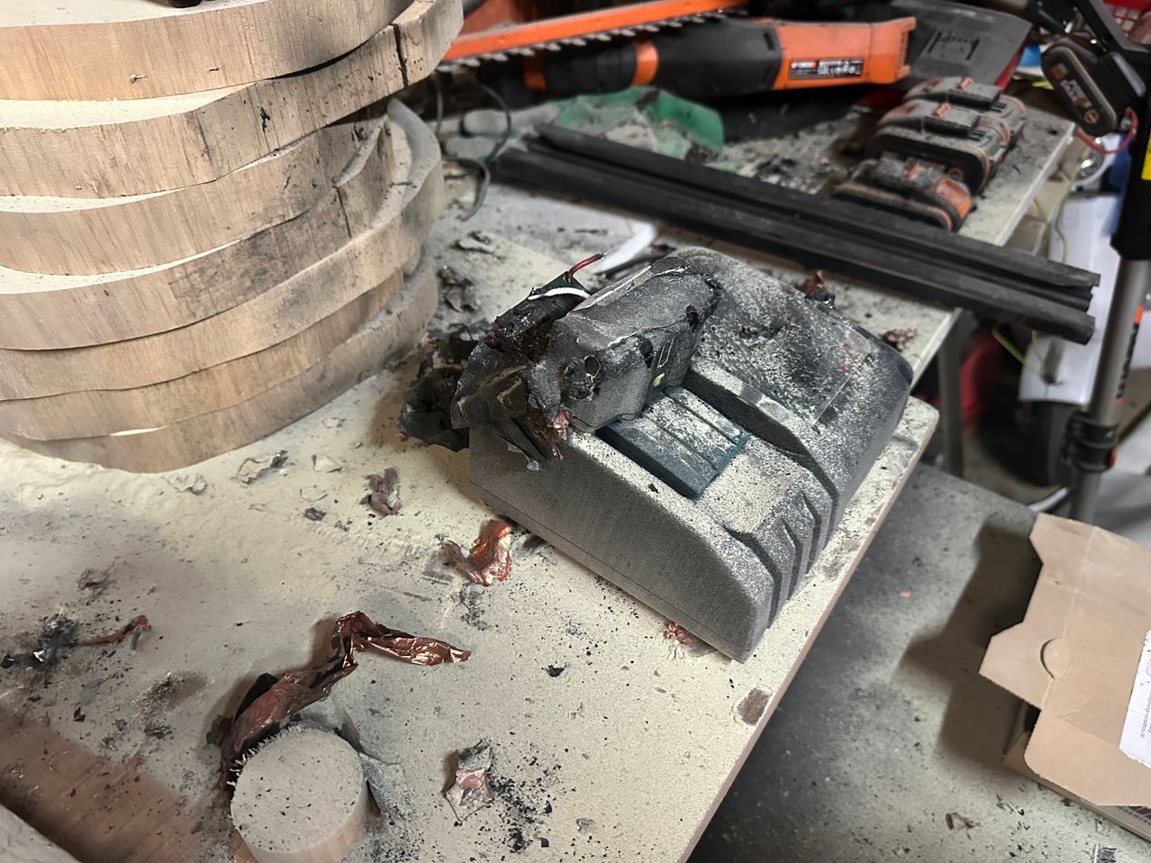 Ein verbranntes Elektrowerkzeug liegt auf einem überfüllten Arbeitsplatz mit Holzbrettern, Metallwerkzeugen und herumliegenden Trümmern.