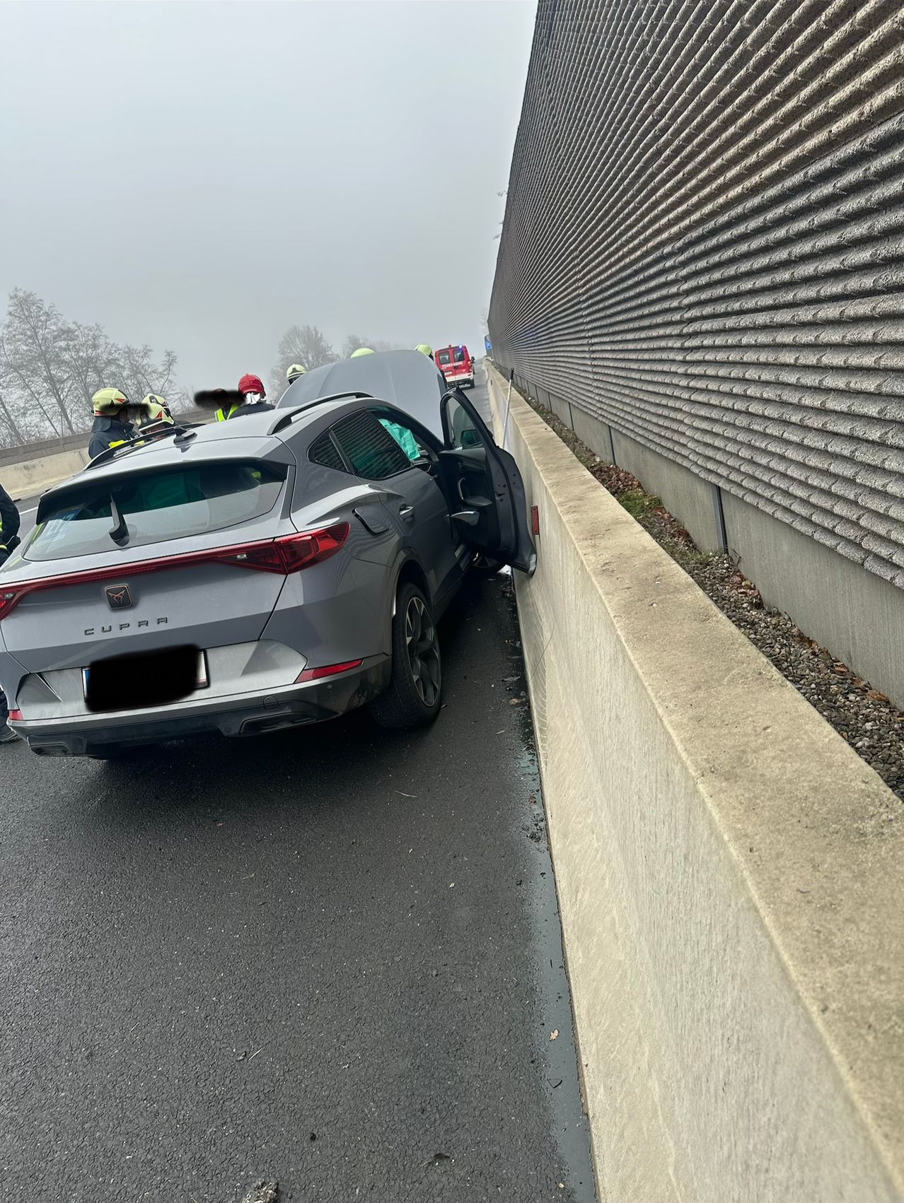 Ein silberner Cupra SUV ist mit einer Betonbarriere auf einer Straße kollidiert. Die Rückseite des Autos ist beschädigt und die hintere Tür ist offen. Rettungskräfte sind anwesend.