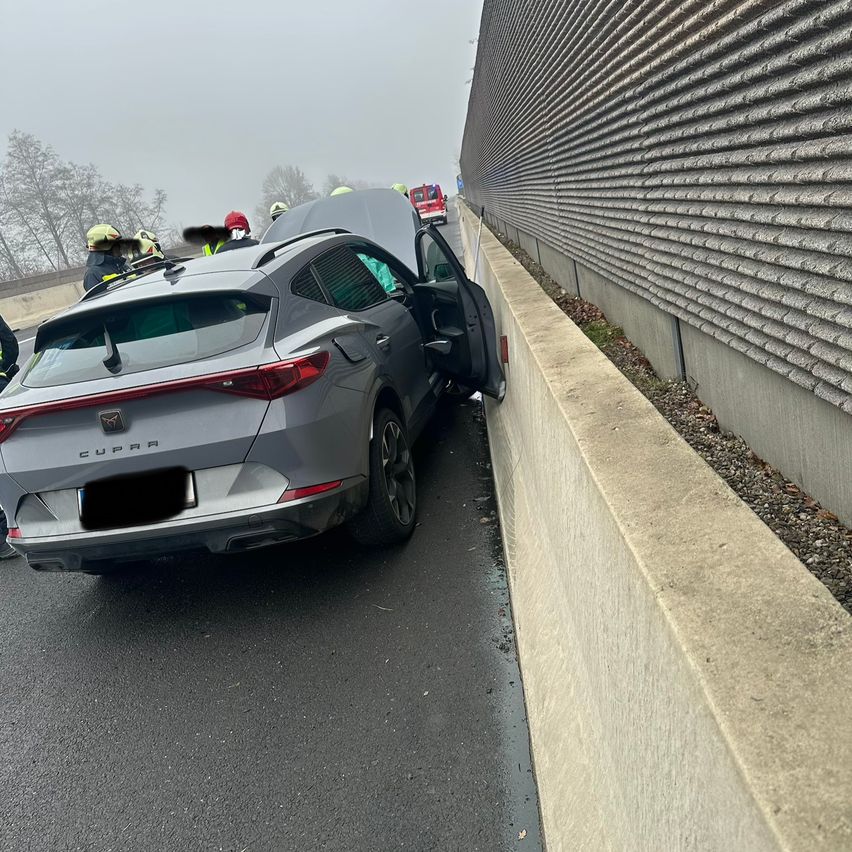 Ein silberner Cupra SUV ist mit einer Betonbarriere auf einer Straße kollidiert. Die Rückseite des Autos ist beschädigt und die hintere Tür ist offen. Rettungskräfte sind anwesend.