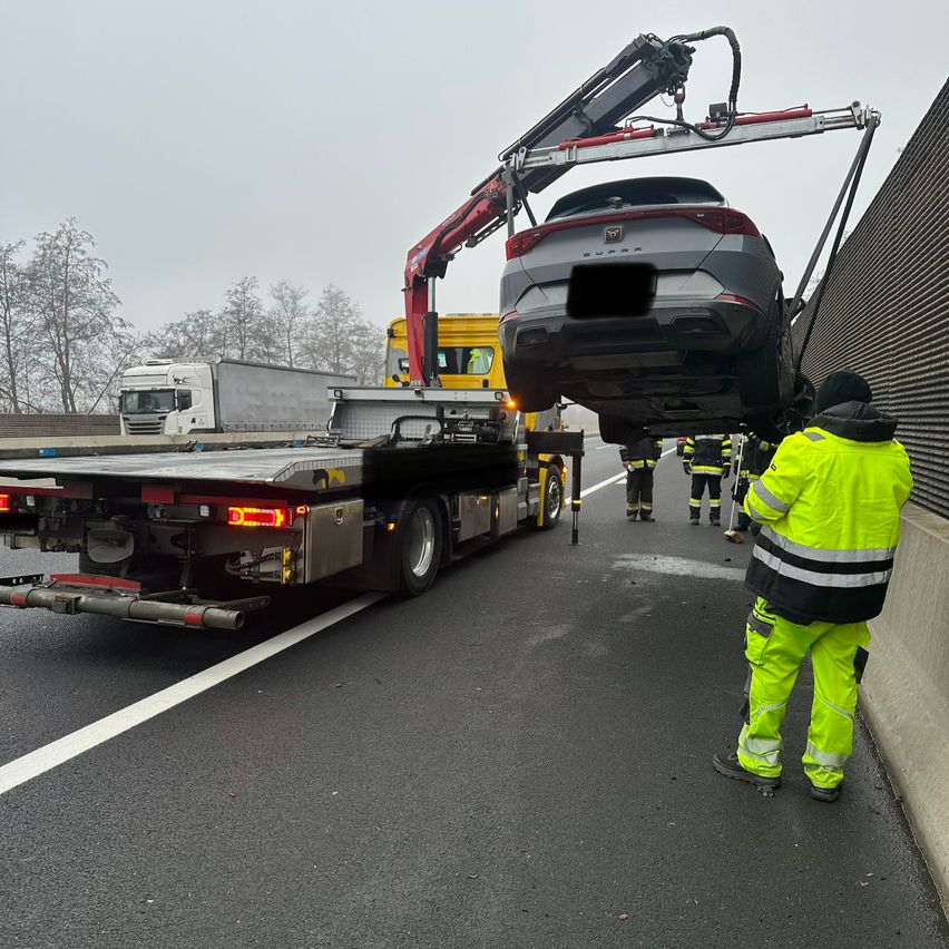 Ein graues Auto wird von einem Kran von der Straße gehoben. Eine Person in einem gelben Mantel steht in der Nähe der Wand. Hinter dem Kran befinden sich mehrere LKWs.