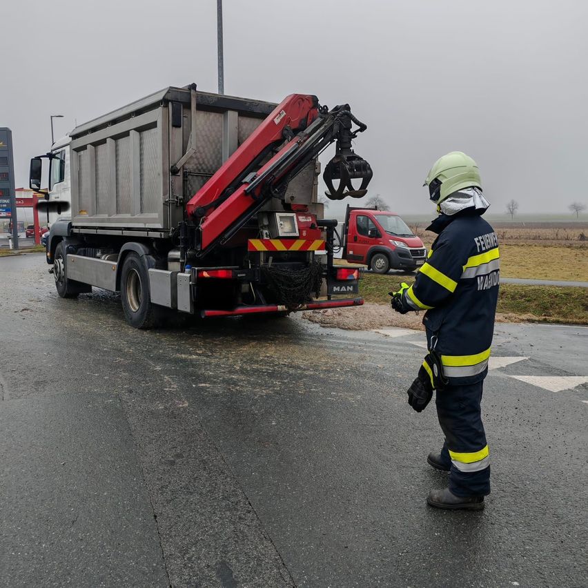 Ein Feuerwehrmann steht neben einem LKW mit einem Kran in einem Außenbereich, möglicherweise an einer Baustelle. Der Kran ist am LKW befestigt.