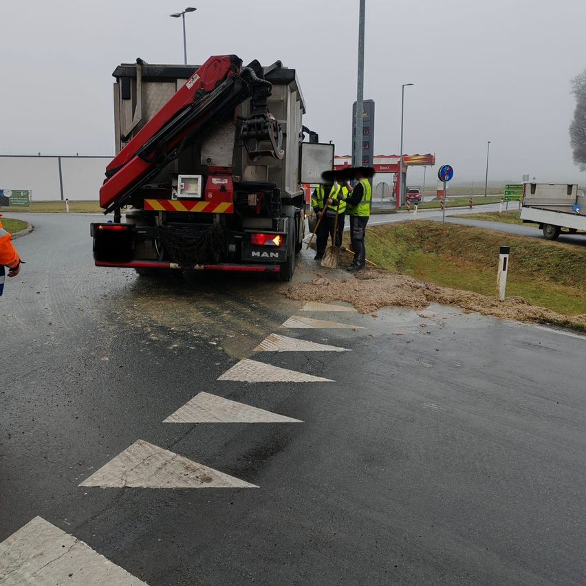 Zwei Arbeiter in gelben Warnwesten fegen neben einem großen Fahrzeug auf der Straße auf. Im verwackelten Hintergrund sind eine Tankstelle, eine Straße und Bäume zu sehen.