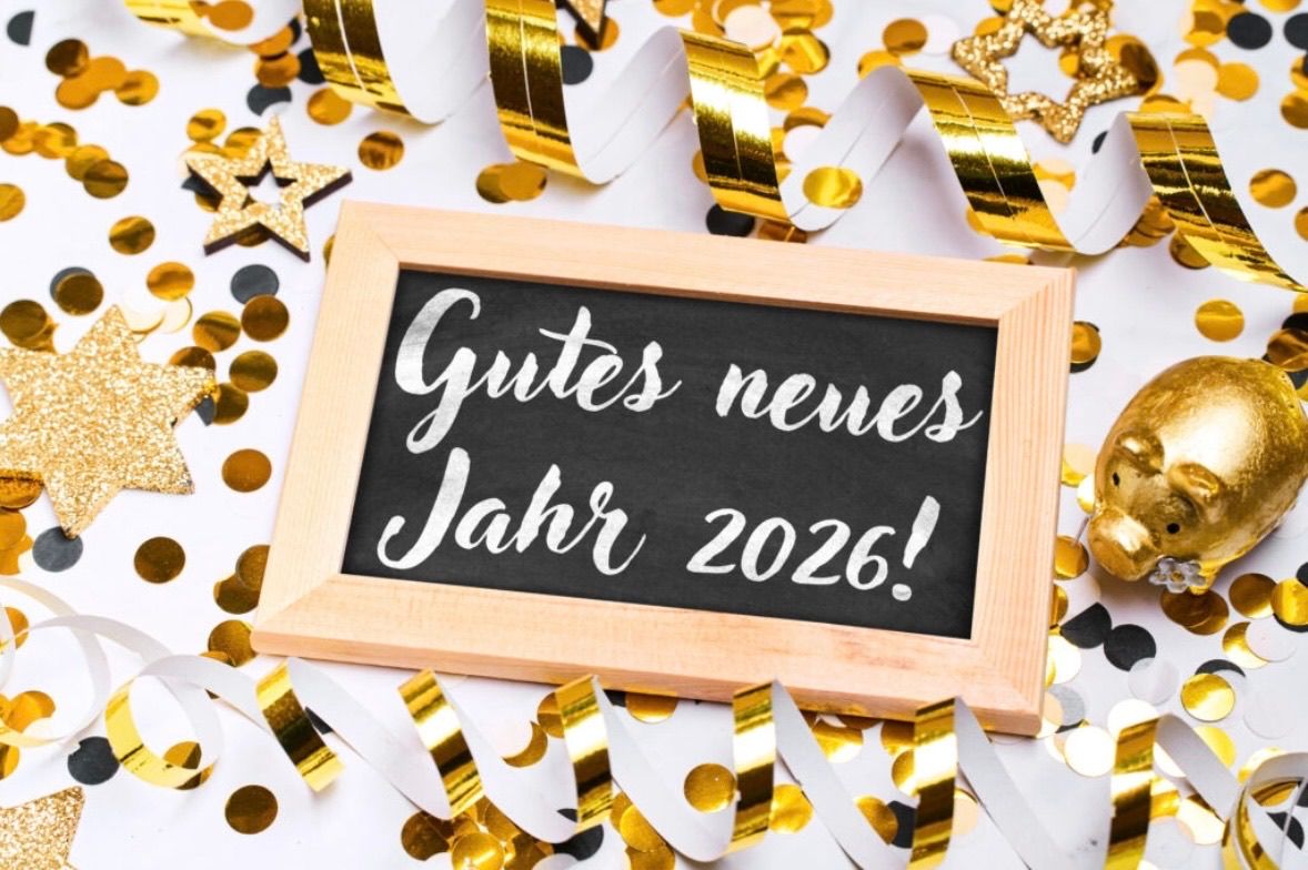 Ein gerahmtes Kreidetafel mit der Botschaft 'Gutes neues Jahr 2026!' ist von goldenen Konfetti und Bändern umgeben.