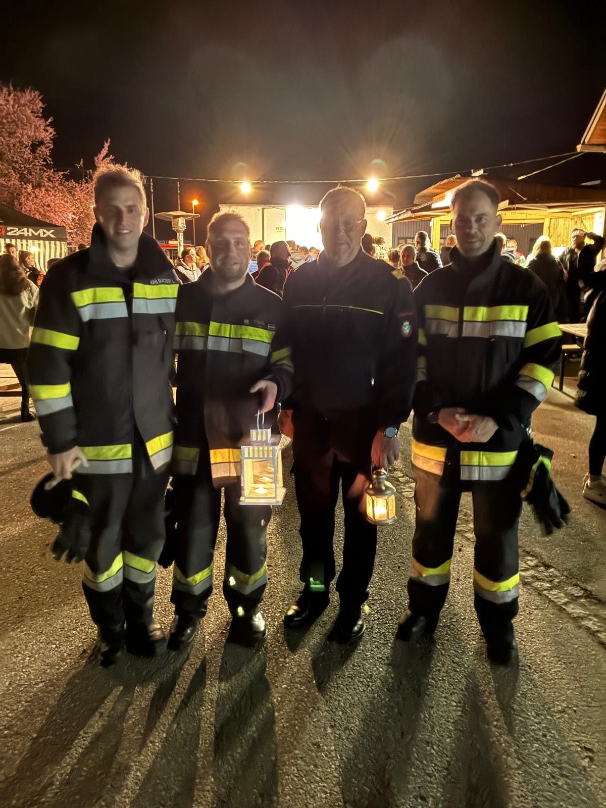 Vier Feuerwehrleute stehen bei Nacht, einer hält eine Laterne. Sie tragen alle einheitliche Uniformen mit reflektierenden Streifen. Der Hintergrund zeigt ein geschäftiges Gebiet mit Menschen und einigen Gebäuden.