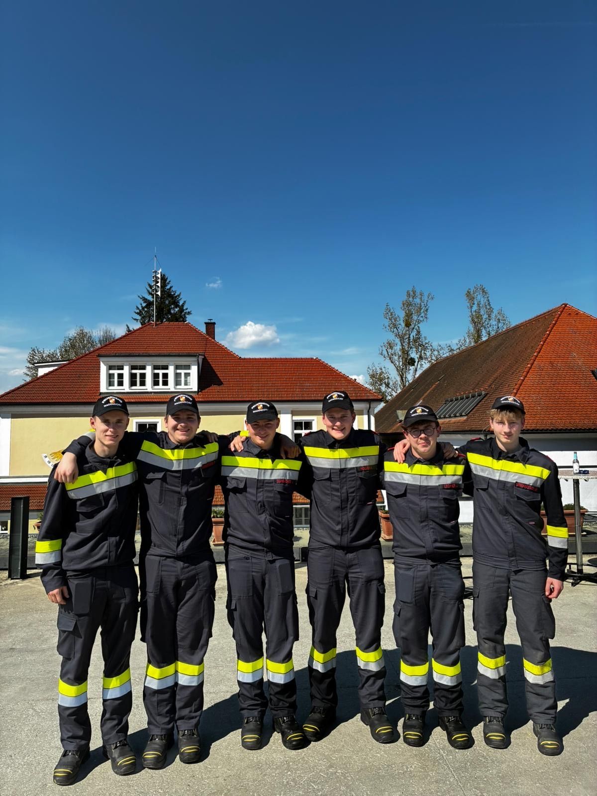 Fünf Feuerwehrleute in Uniform stehen vor einem Haus mit roten Dächern und klarem blauem Himmel. Bäume und ein klarer Himmel sind hinter ihnen.