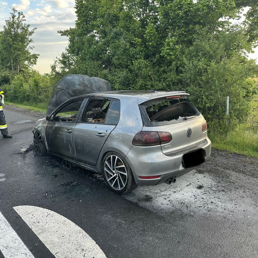 Ein beschädigter silberner Volkswagen Golf steht am Straßenrand, mit einem Felsbrocken auf dem Dach. Ein Feuerwehrmann ist in der Nähe, und das Gebiet ist von Grün umgeben.