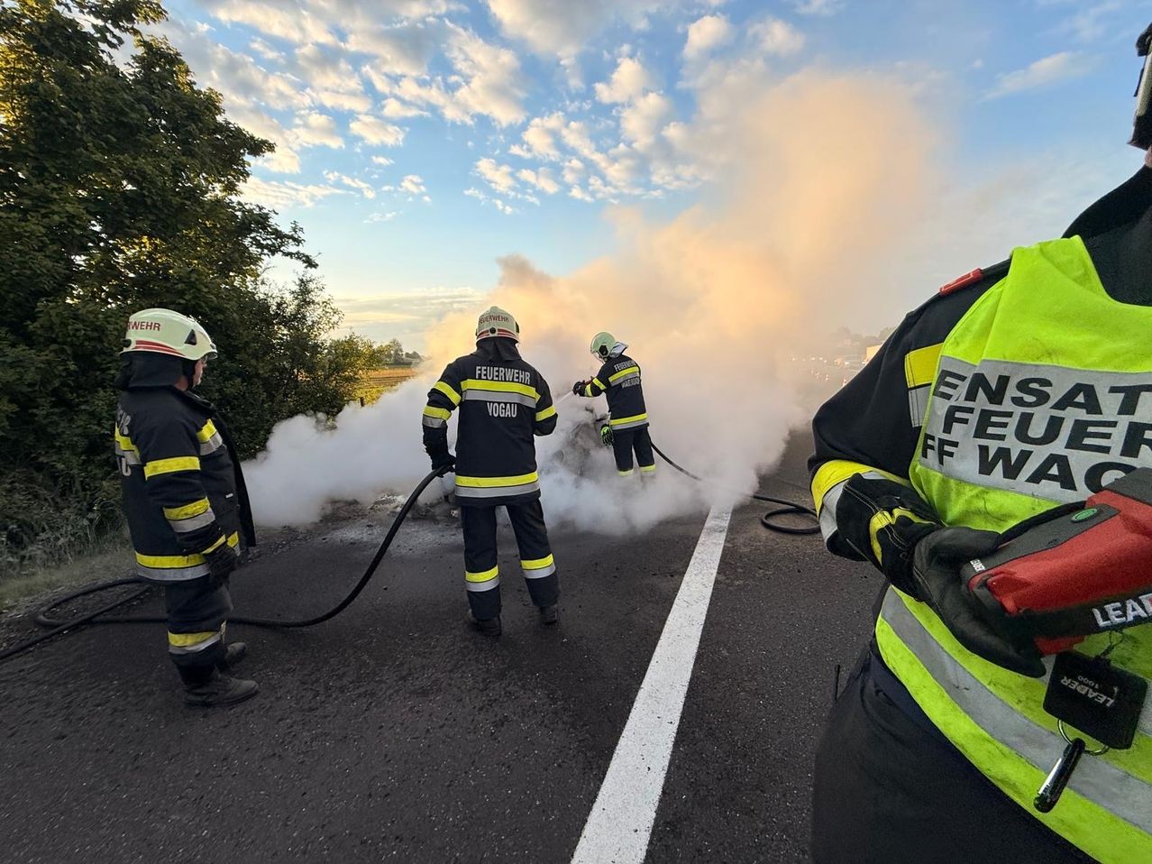 Feuerwehrleute in Uniformen sind auf einer Straße und löschen ein Feuer mit einem Schlauch. Rauch steigt vom brennenden Objekt auf.