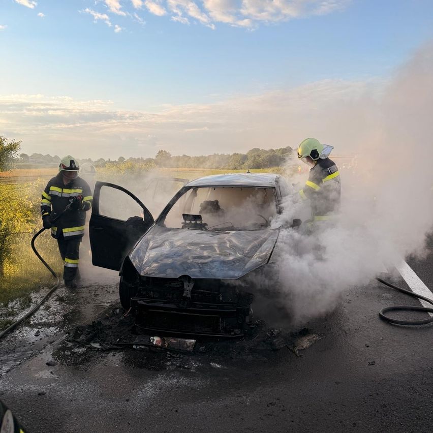 Feuerwehrleute sind an einer Straße mit einem brennenden Auto. Einer spritzt Wasser, während der andere zusieht. Rauch steigt vom Auto und der Umgebung auf.