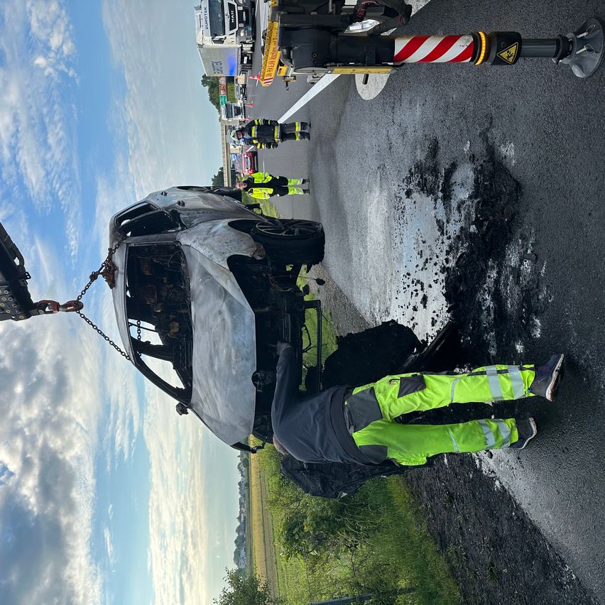 Ein schwer beschädigtes Auto wird auf einer Straße von einem Kran angehoben, wobei Feuerwehrleute und Rettungsfahrzeuge anwesend sind.