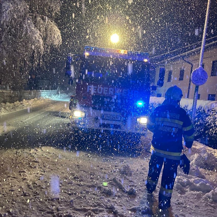 Ein Feuerwehrmann steht im Schnee neben einem Feuerwehrwagen mit blinkenden Lichtern auf einer verschneiten Straße in der Nacht.