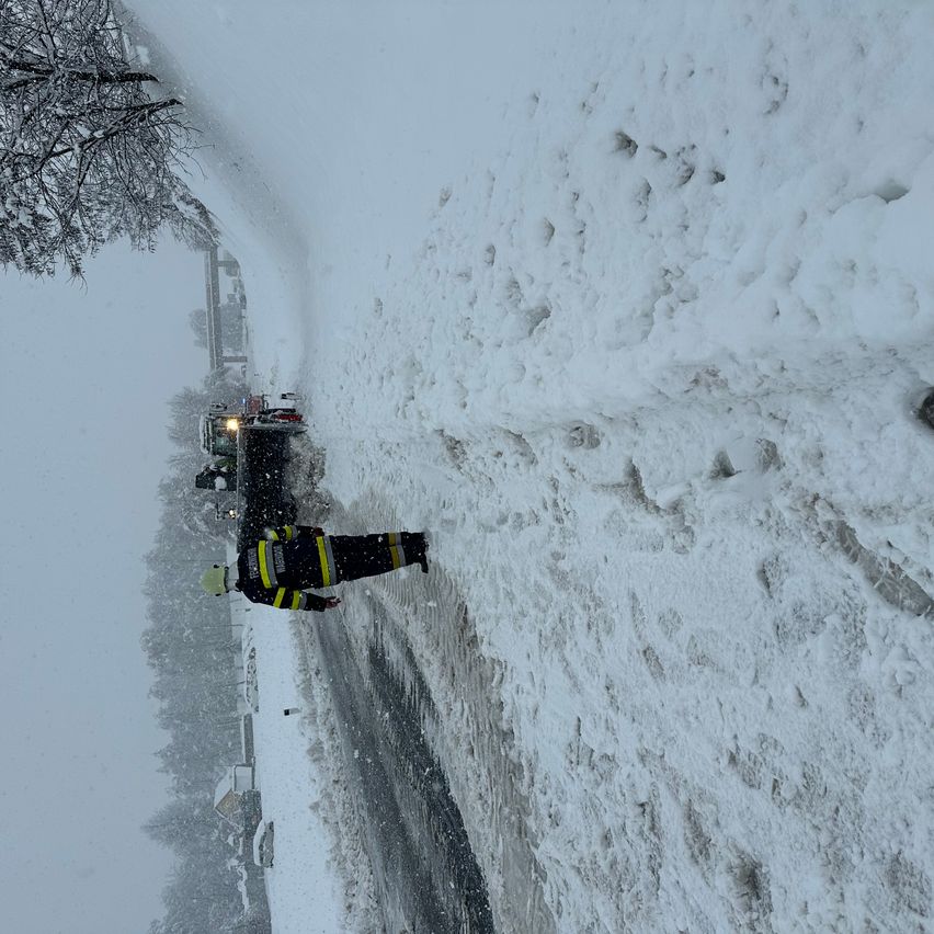 Ein Feuerwehrmann in Uniform steht auf einer verschneiten Straße, mit einem Schneepflugfahrzeug hinter ihm. Schneebedeckte Bäume und ein Gebäude sind auf der linken Seite zu sehen.