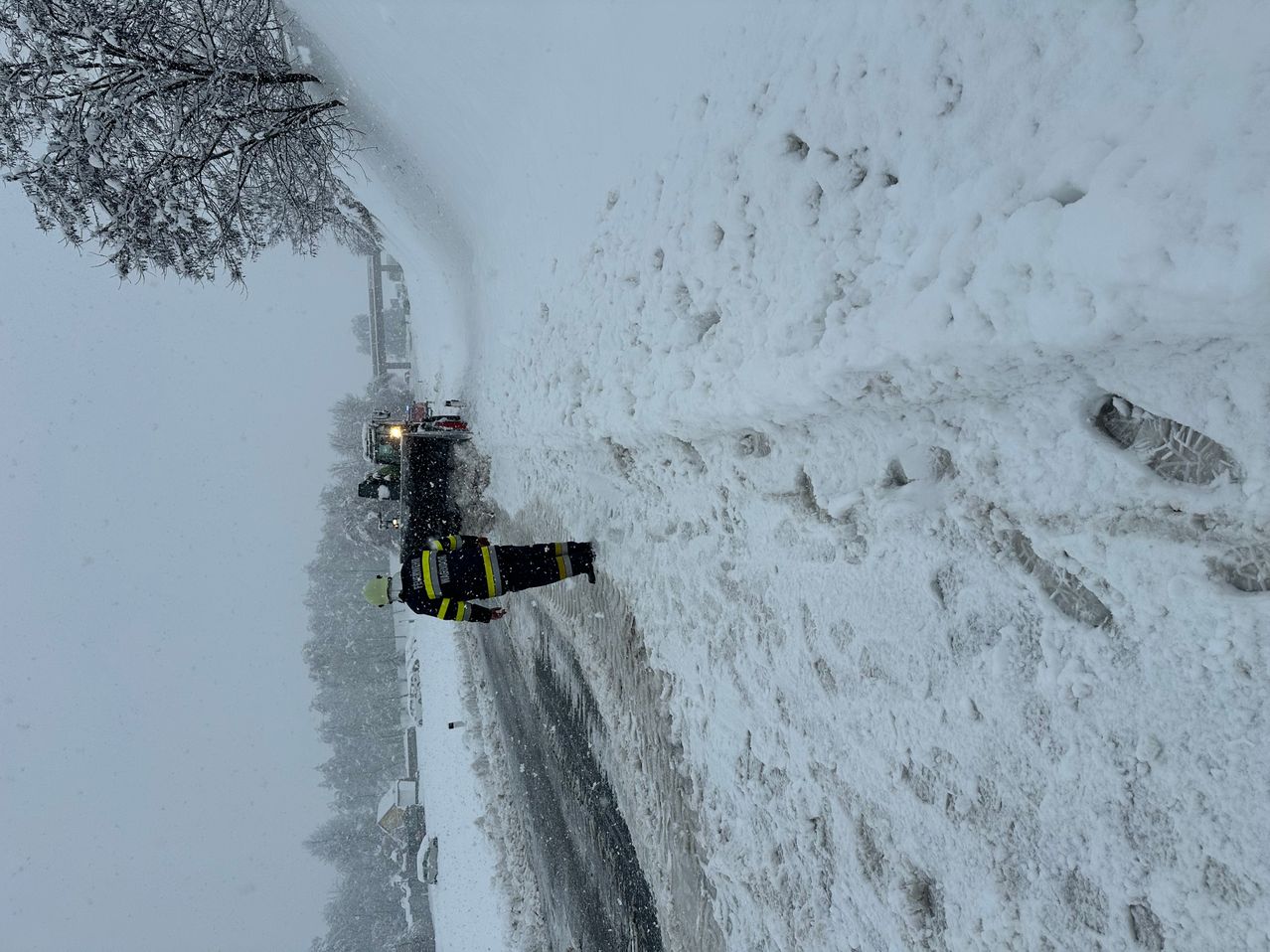 Ein Feuerwehrmann in Uniform steht auf einer verschneiten Straße, mit einem Schneepflugfahrzeug hinter ihm. Schneebedeckte Bäume und ein Gebäude sind auf der linken Seite zu sehen.