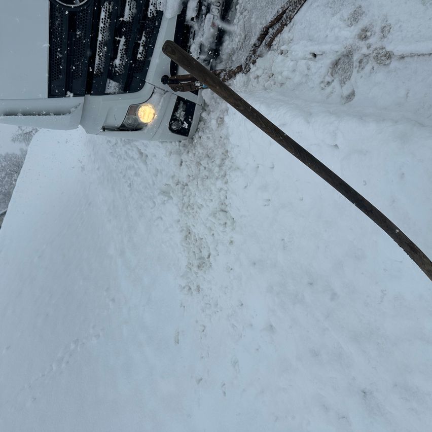 Ein Fahrzeug ist teilweise im Schnee vergraben, die Scheinwerfer sind eingeschaltet. Ein Holzstab ist vor dem Fahrzeug platziert.