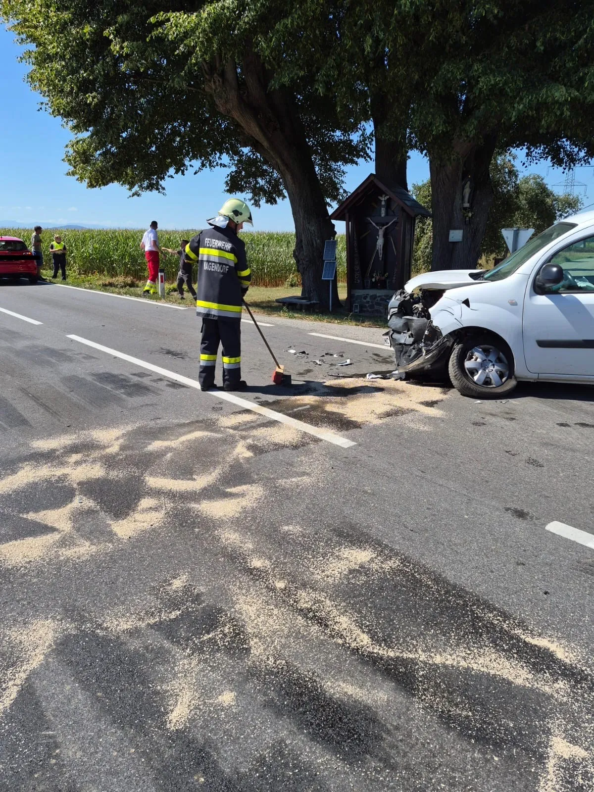 Ein Feuerwehrmann reinigt Trümmer von einem Verkehrsunfall mit einem weißen Van. In der Nähe sind eine kleine Kapelle und ein Maisfeld zu sehen.