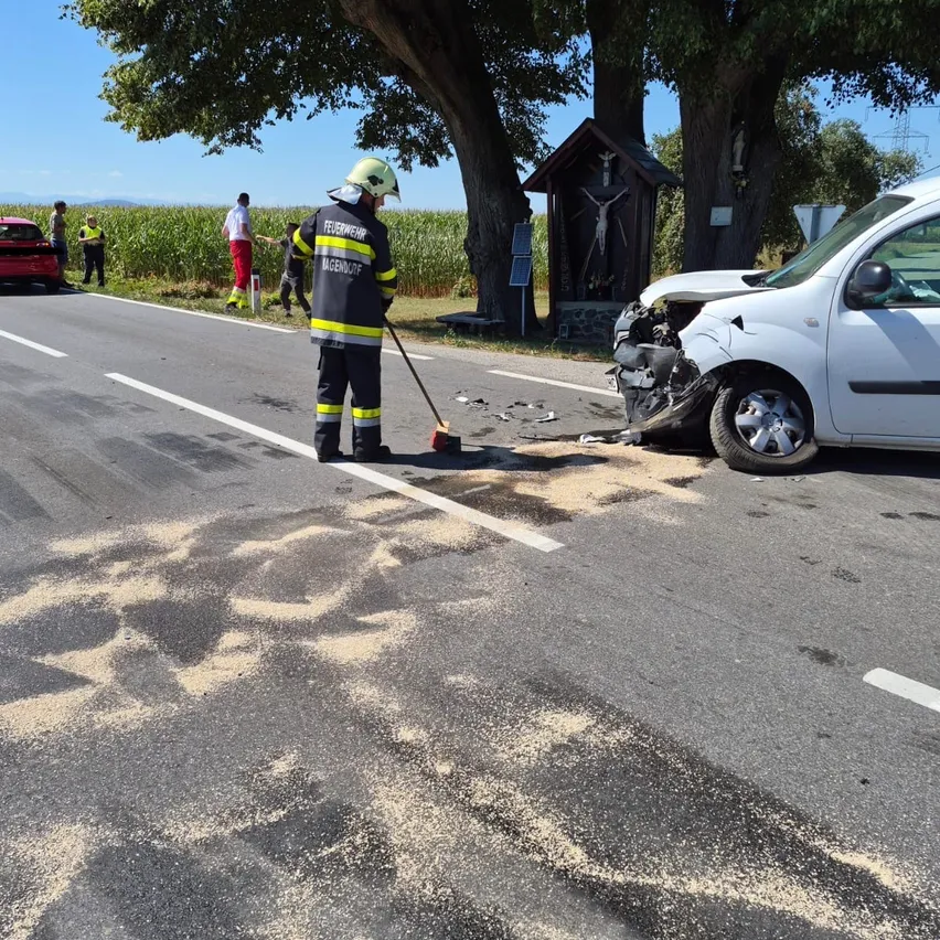Ein Feuerwehrmann reinigt Trümmer von einem Verkehrsunfall mit einem weißen Van. In der Nähe sind eine kleine Kapelle und ein Maisfeld zu sehen.