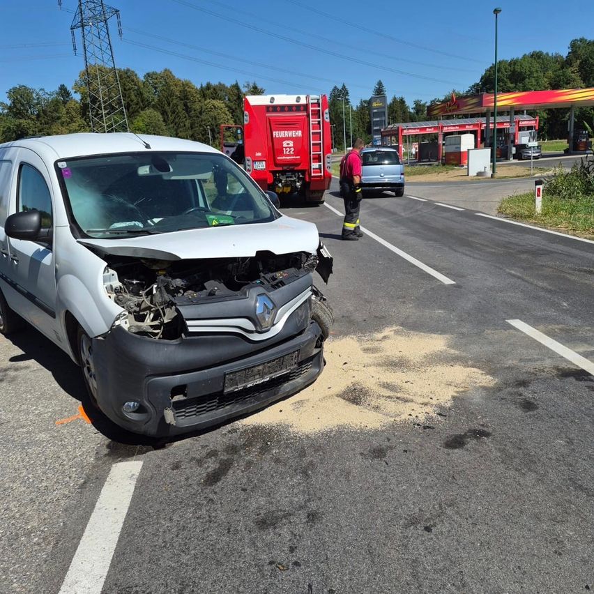 Ein weißer Renault-Transporter mit beschädigter Front ist am Straßenrand geparkt. Ein Feuerwehrmann steht in der Nähe, und ein roter Feuerwehrwagen steht hinter dem Van.