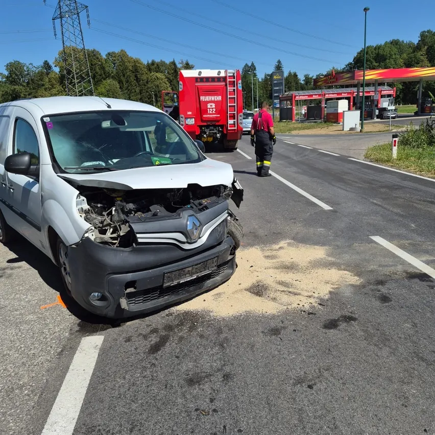 Ein weißer Lieferwagen mit zertrümmerter Front ist auf der Straße geparkt, mit einem Feuerwehrwagen und einer Person in Schutzausrüstung in der Nähe.