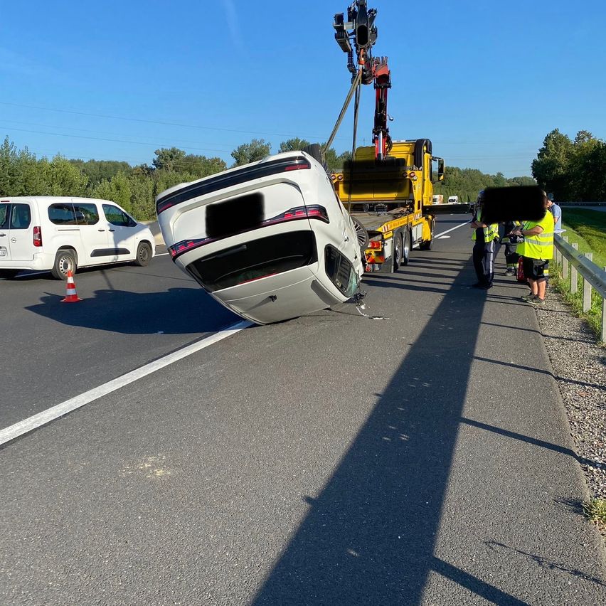 Ein weißer Wagen liegt auf der Seite an der Straßenseite, ein Abschleppwagen hebt ihn an. Ein weißer Van und Verkehrskegel sind in der Nähe.