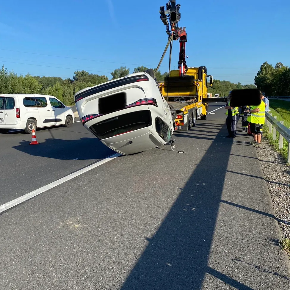 Ein weißer Wagen liegt auf der Seite an der Straßenseite, ein Abschleppwagen hebt ihn an. Ein weißer Van und Verkehrskegel sind in der Nähe.