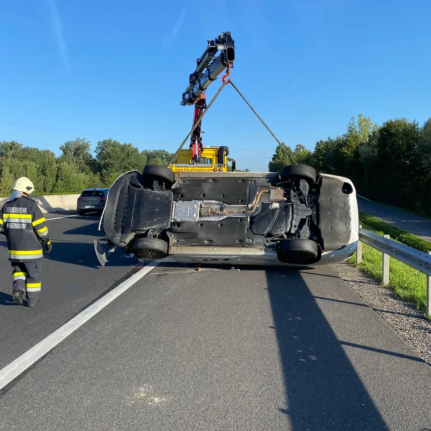 Ein Feuerwehrmann steht neben einer Straße, wo ein Kran ein umgestürztes Fahrzeug hebt. Die Szene spielt sich an einem sonnigen Tag mit Bäumen und Strommasten im Hintergrund ab.