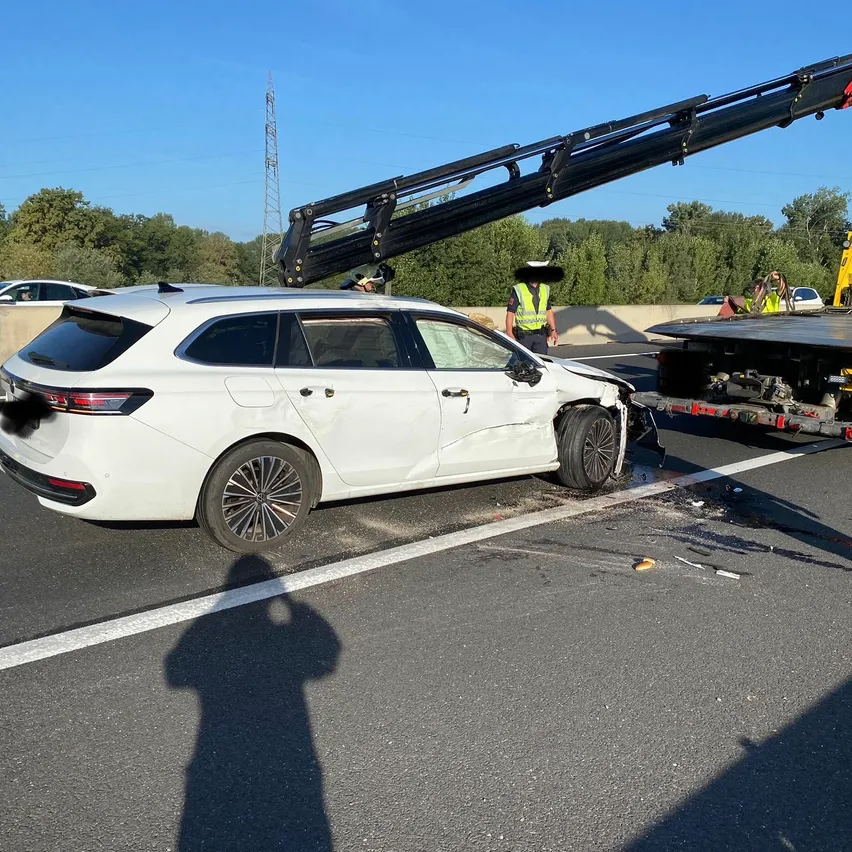 Ein weißer SUV wird von einem Kran auf einer Autobahn abgeschleppt. Das Fahrzeug hat Schäden am Heck erlitten. Eine Person in einer reflektierenden Weste steht in der Nähe.
