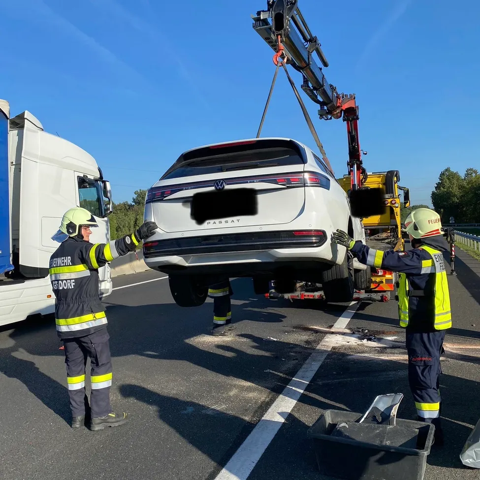 Rettungskräfte entfernen einen beschädigten weißen Volkswagen Passat von der Straße mit einem Kran. Das Fahrzeug wird von hinten angehoben, und zwei Arbeiter stehen auf beiden Seiten.