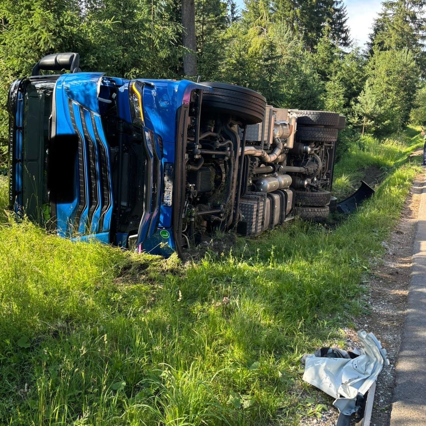 Ein blauer Lkw liegt auf der Seite am Straßenrand, mit sichtbaren Reifen und Auspuff. In der Nähe befindet sich ein Stück zerrissenes Plastik.