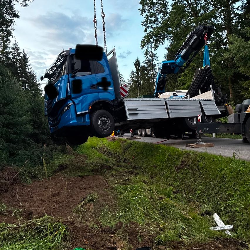 Ein blauer Lastwagen wird von einem Kran von der Straßenseite angehoben, in der Nähe einiger Bäume.