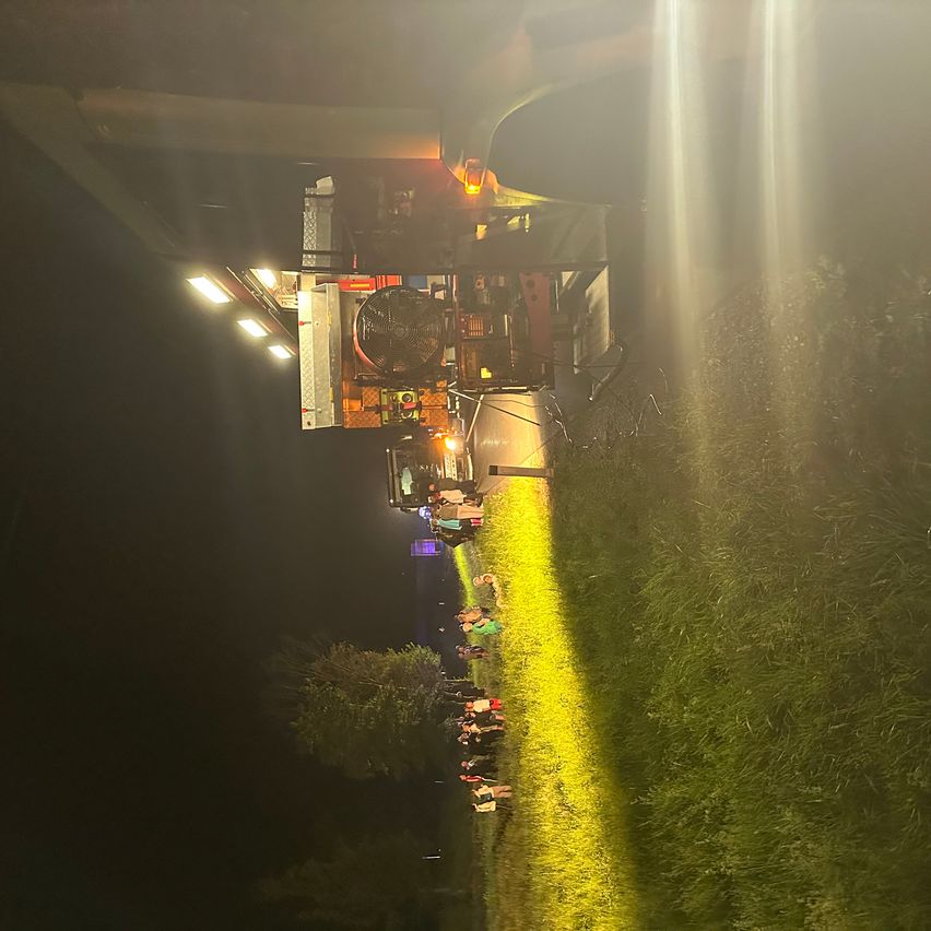 Bei Nacht steht ein Fahrzeug mit gelbem Licht am Straßenrand, mit Menschen, die auf dem Rasen stehen.