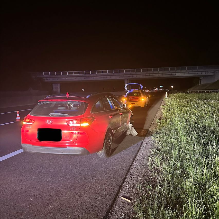 Ein rotes Auto mit einem beschädigten Stoßfänger ist nachts am Straßenrand geparkt. Ein weiteres Fahrzeug mit offenem Kofferraum befindet sich ebenfalls in der Nähe.