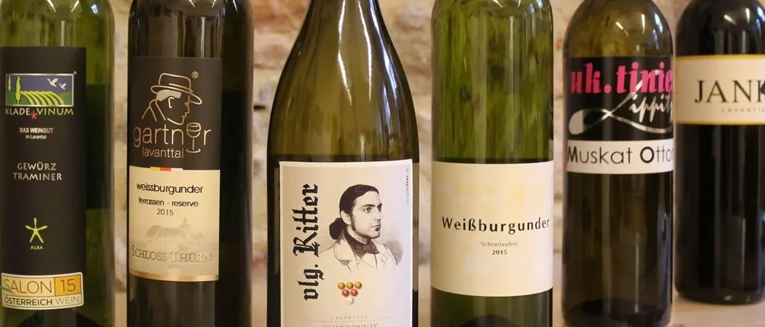 Eine Reihe von sechs Weinflaschen mit verschiedenen Etiketten. Die dritte Flasche von links hat ein Bild eines Mannes auf ihrem Etikett. Hinter den Flaschen ist eine Steinmauer.