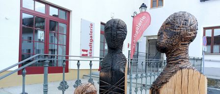 Drei Holzfiguren stehen vor einem weißen Gebäude mit roten Rahmen und einer roten Fahne mit der Aufschrift 'stadtgalerie'.