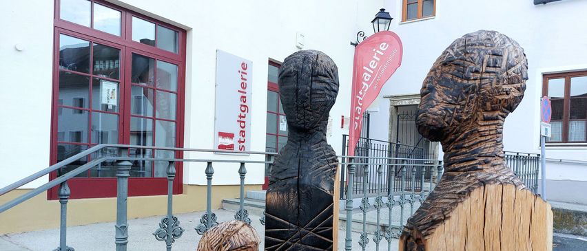 Drei Holzfiguren stehen vor einem weißen Gebäude mit roten Rahmen und einer roten Fahne mit der Aufschrift 'stadtgalerie'.