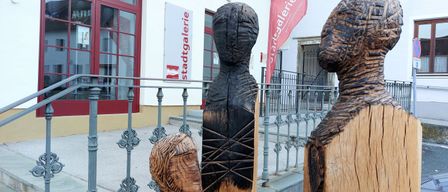 Drei Holzskulpturen sind vor einem Gebäude mit einem roten Banner ausgestellt. Die Skulpturen stehen vor einer weißen Wand mit roten Rahmen.
