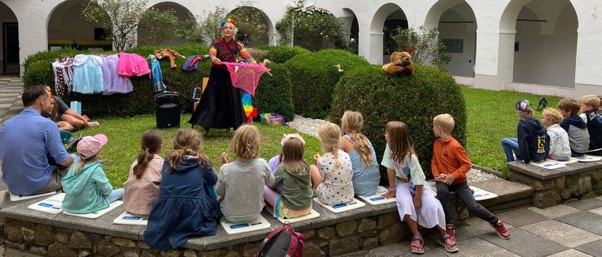 Eine Frau unterhält eine Gruppe von Kindern, die auf einer Steinbank draußen vor einem weißen Gebäude sitzen.