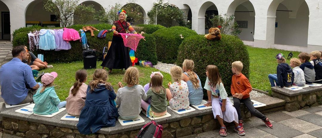 Eine Frau unterhält eine Gruppe von Kindern, die auf einer Steinbank draußen vor einem weißen Gebäude sitzen.
