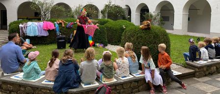 Ein Künstler unterhält eine Gruppe von Kindern vor einem weißen Gebäude mit Rundbogenfenstern.