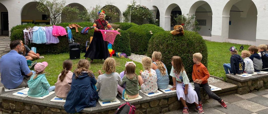 Ein Künstler unterhält eine Gruppe von Kindern vor einem weißen Gebäude mit Rundbogenfenstern.